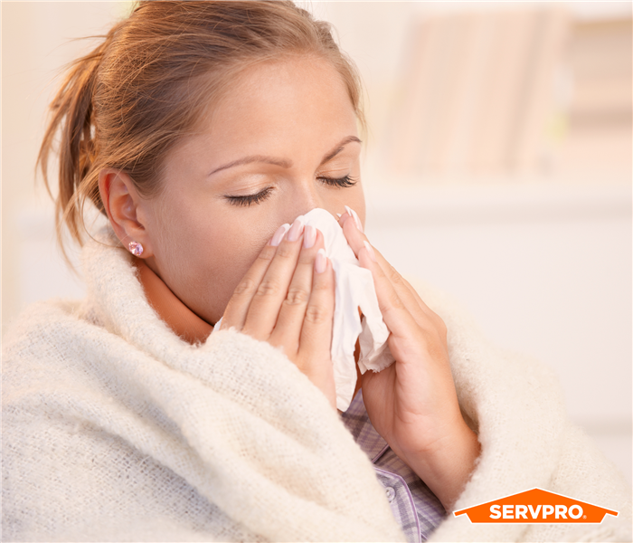 woman with hair up, blowing nose into a tissue in a bathrobe, orange servpro logo in bottom right