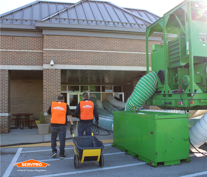 Two SERVPRO employees in orange vests walking into a school next to a big, green desiccant dehumidifier 